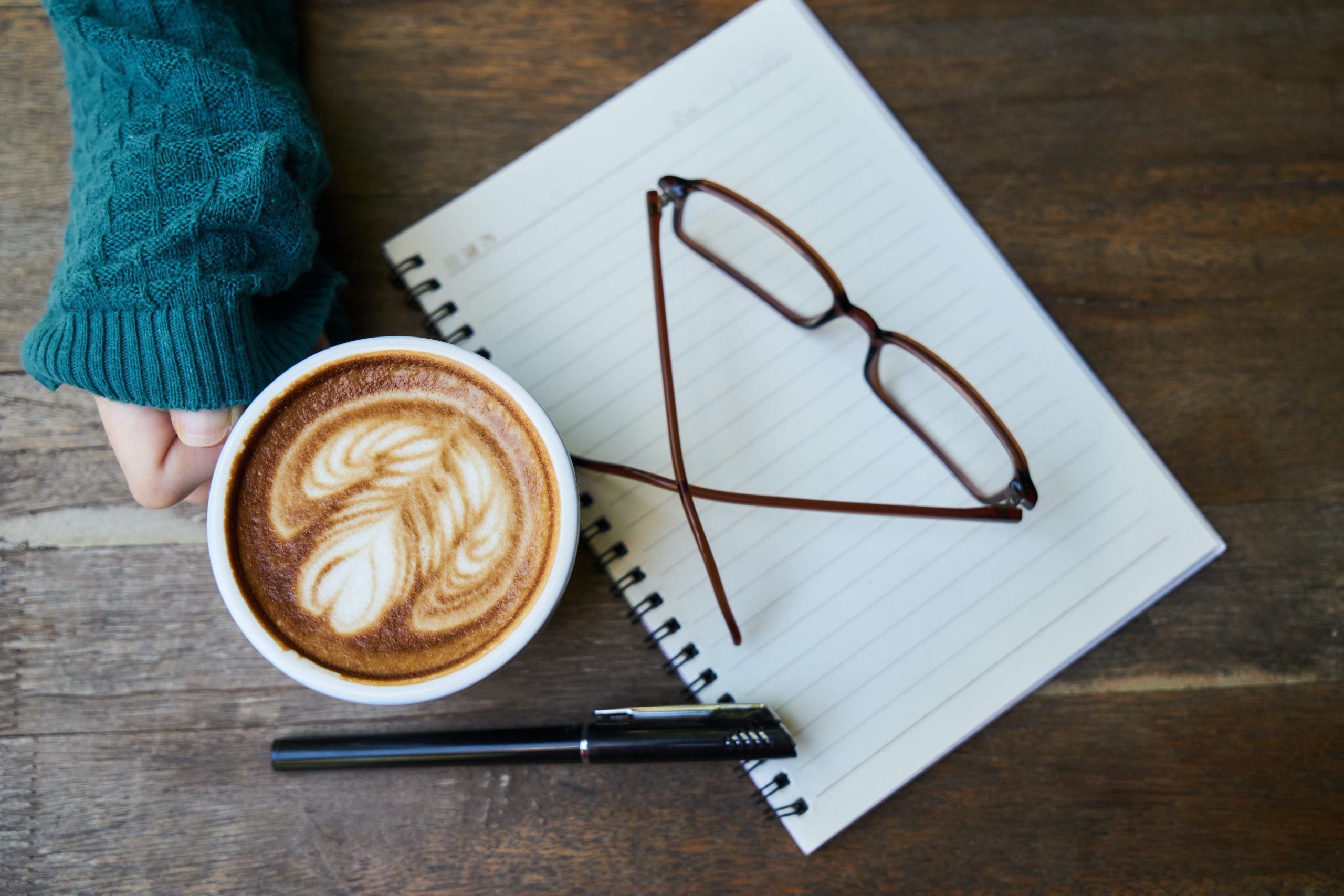 Kaffekopp, glasögon, skrivblock och penna på ett träbord