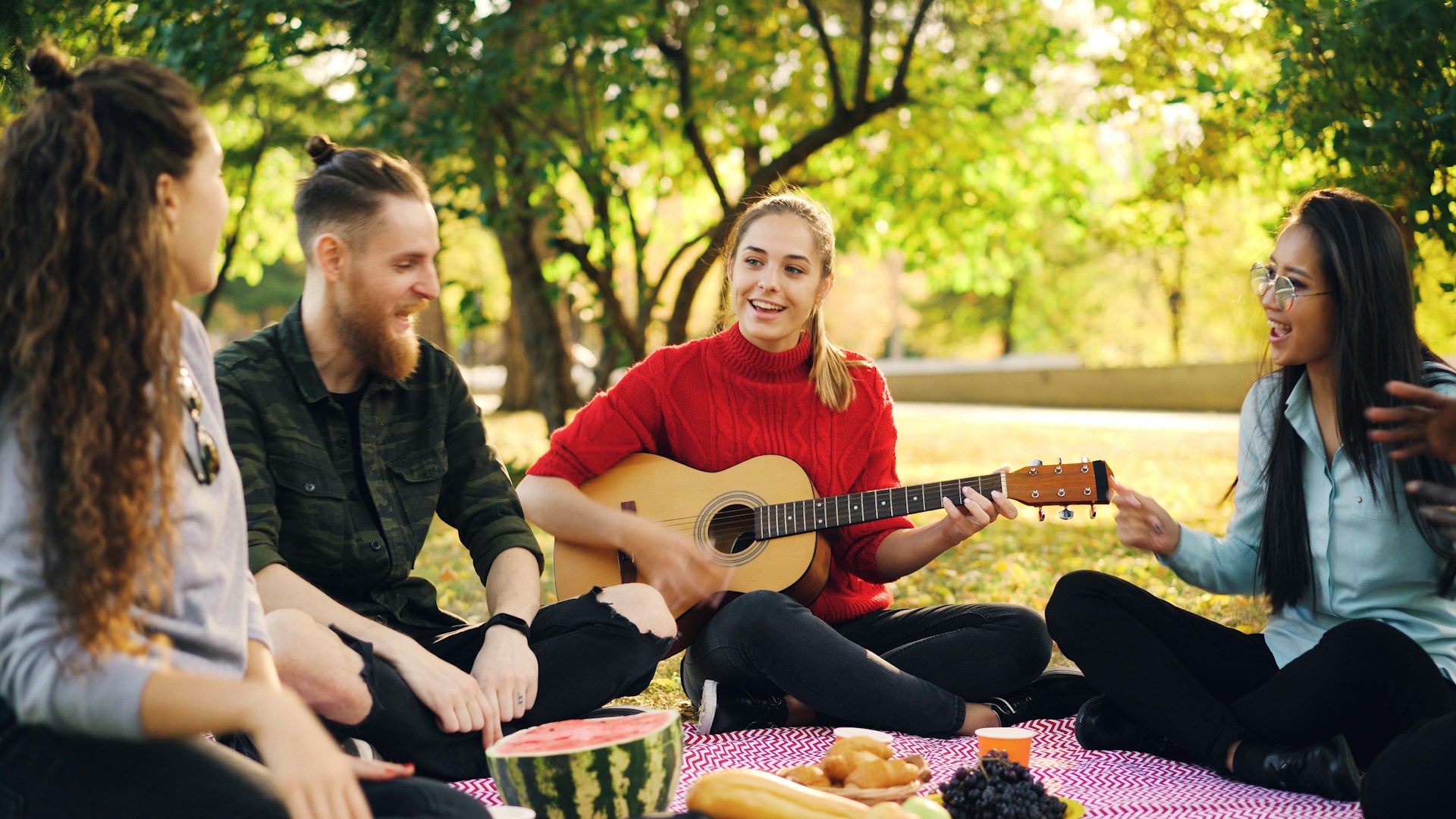 Bild på fyra personer som sitter tillsammans på en filt utomhus, en spelar gitarr.