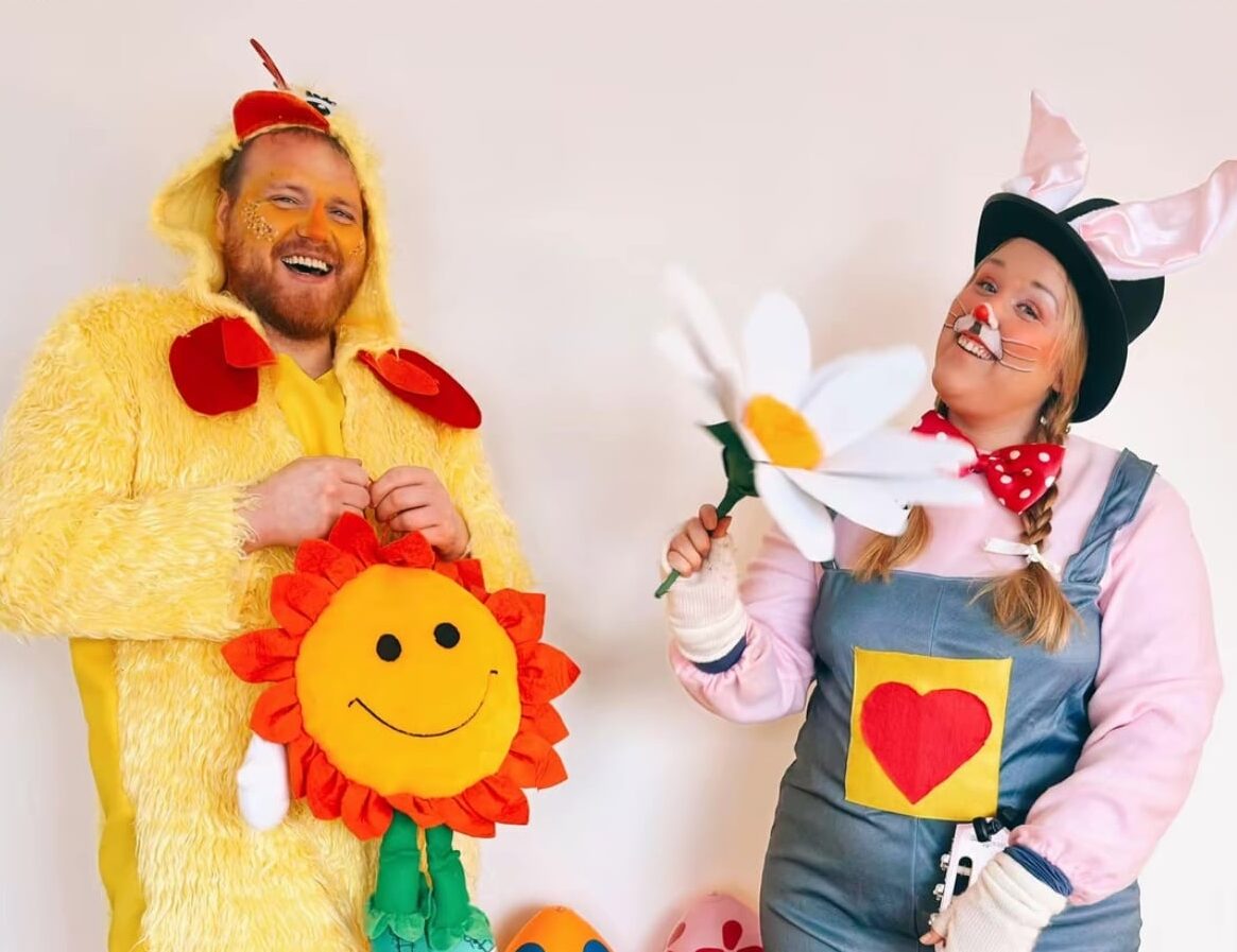En glad rödskäggig man utklädd till gul kyckling som håller i en gul och röd stor goseblomma och en glad kvinna utklädd till rosa kanin med hängselbyxor och som håller en stor vit och gul tygblomma.