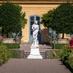 Bild på staty i Linnéträdgården, man ser även Linnéhuset i bakgrunden och växter runtomkring.