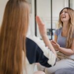 Två flickor sitter i en grå soffa, pratar och skrattar.