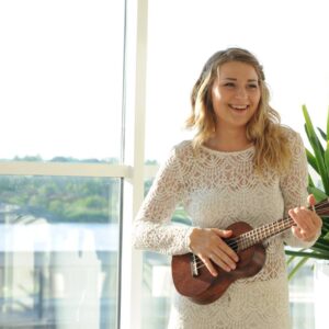 Kvinna i vit klänning med ukulele framför fönster.