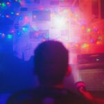 Bilden visar en person sedd bakifrån i ett mörkt rum, upplyst av starka röda och blå ljus. Det är oklart exakt vad personen tittar på, men det är en vägg full av olika tidningspapper och färgglada lampor.