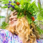 En flicka som bär en blomsterkrans på huvudet med gröna blad och färgglada blommor. Hon har långt, ljust hår och ler mjukt. hon har ögonen slutna medan sommar solen skiner.