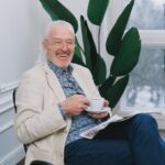 En skrattande äldre man som håller i en kaffekopp och har en tidning i knät.