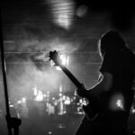 En svartvit bild på en långhårig man som syns snett bakifrån och spelar på en elgitarr på en scen.