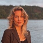 Clara Alm inför Musik i sommarkväll i Husby-Långhundra kyrka, med kvällsljus vid vattnet i bakgrunden.