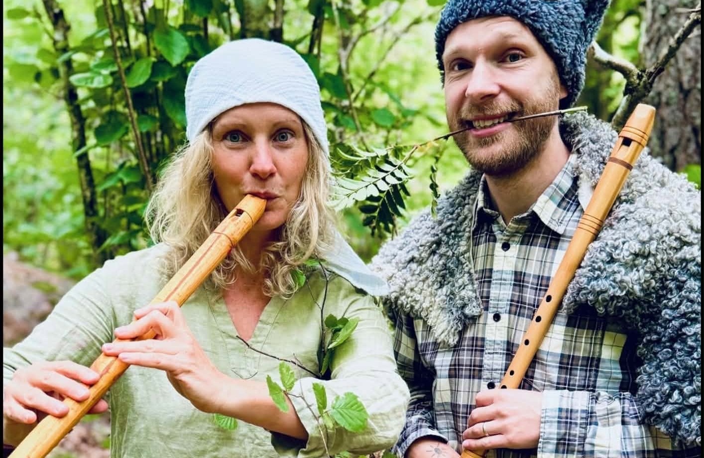 Halvbild på en kvinna och en man i skogen med varsin flöjt. Mannen har en rönnkvist i munnen.