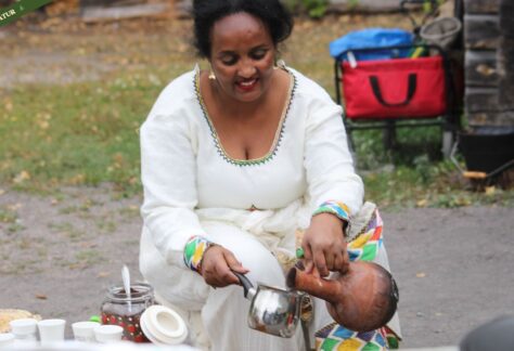 En eritreansk kvinna sitter utomhus och häller upp en dryck. Hon är klädd i en vit traditionell klänning med färgglada detaljer runt halsen och ärmarna.