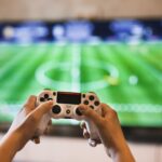 En person som spelar Fifa, man ser fotbollsplanen på TV:n och två händer som håller i en kontroll.