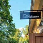 Ingången till ett gult äldre hus på höger sida med en skylt där det står "UPPSALA FÖRFATTARSÄLLSKAP". Lövverk från träd på vänstra sidan.