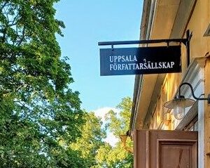 Ingången till ett gult äldre hus på höger sida med en skylt där det står "UPPSALA FÖRFATTARSÄLLSKAP". Lövverk från träd på vänstra sidan.