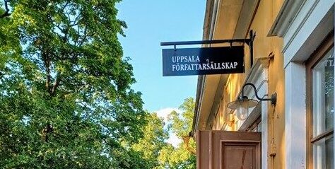 Ingången till ett gult äldre hus på höger sida med en skylt där det står "UPPSALA FÖRFATTARSÄLLSKAP". Lövverk från träd på vänstra sidan.