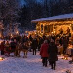 Julmarknad i vinterlandskap. En julpyntad dansbana på höger sida med två kvinnor som spelar musik. Försäljningsstånd, människor som går runt, barn som åker häst och vagn, en eldshow, eldkorg och marschaller.