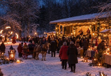 Julmarknad i vinterlandskap. En julpyntad dansbana på höger sida med två kvinnor som spelar musik. Försäljningsstånd, människor som går runt, barn som åker häst och vagn, en eldshow, eldkorg och marschaller.