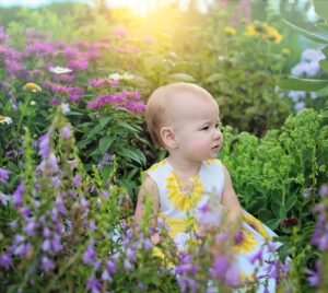Bild på en bebis som sitter ute i naturen omringad av växter och blommor med solljus bakom sig.